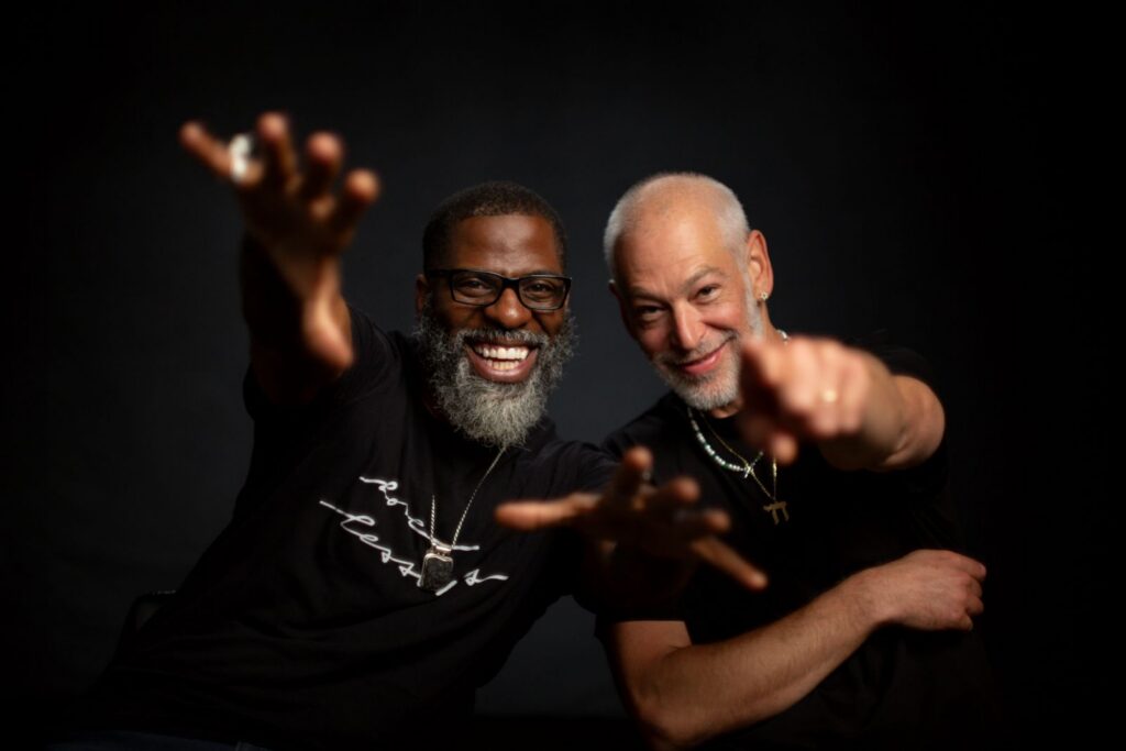 Two men stand together looking excited and pointing at the camera. The man on the left is Black and has glasses and a grey beard. The Man on the right is Jewish and also has a grey beard. They are both smiling and wearing Black t-shirts.