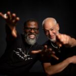 Two men stand together looking excited and pointing at the camera. The man on the left is Black and has glasses and a grey beard. The Man on the right is Jewish and also has a grey beard. They are both smiling and wearing Black t-shirts.