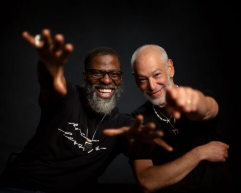 Two men stand together looking excited and pointing at the camera. The man on the left is Black and has glasses and a grey beard. The Man on the right is Jewish and also has a grey beard. They are both smiling and wearing Black t-shirts.
