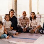 An old photograph shows an Iranian family sitting together on the floor. They are all looking at the camera and smiling.