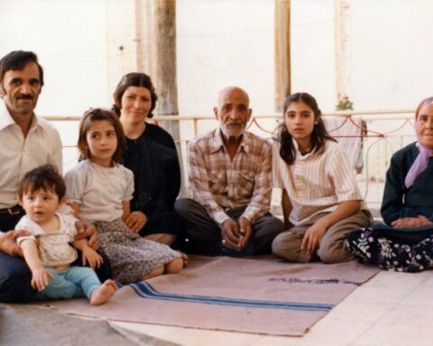 An old photograph shows an Iranian family sitting together on the floor. They are all looking at the camera and smiling.