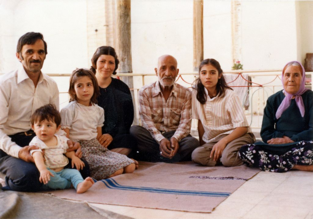 An old photograph shows an Iranian family sitting together on the floor. They are all looking at the camera and smiling.