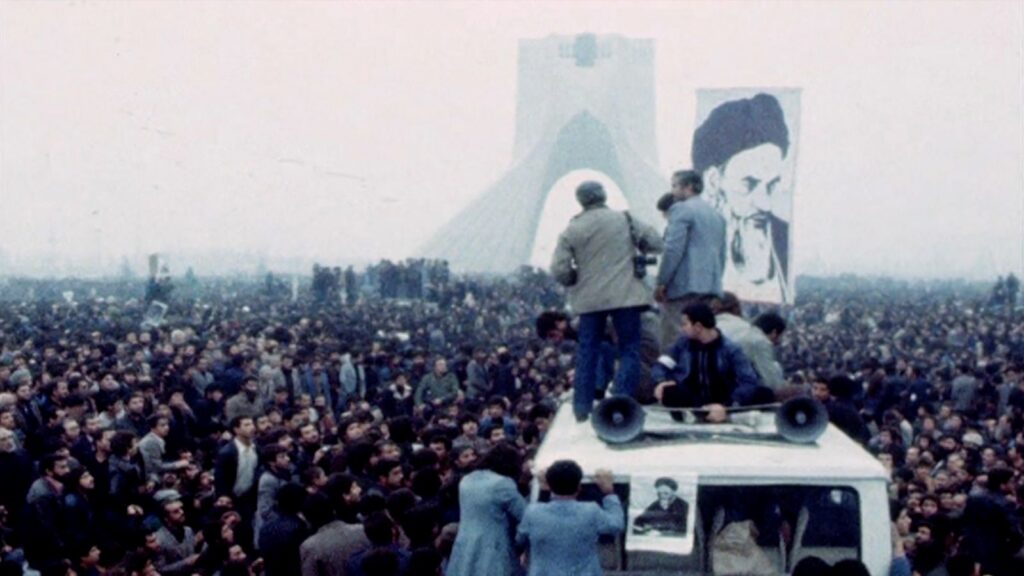 A crowd of people protest in Iran. A few people stand in the foreground, while a large picture of the Shah is in the background.