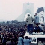 A crowd of people protest in Iran. A few people stand in the foreground, while a large picture of the Shah is in the background.
