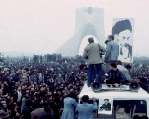 A crowd of people protest in Iran. A few people stand in the foreground, while a large picture of the Shah is in the background.