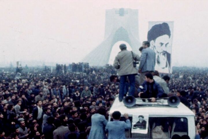 A crowd of people protest in Iran. A few people stand in the foreground, while a large picture of the Shah is in the background.