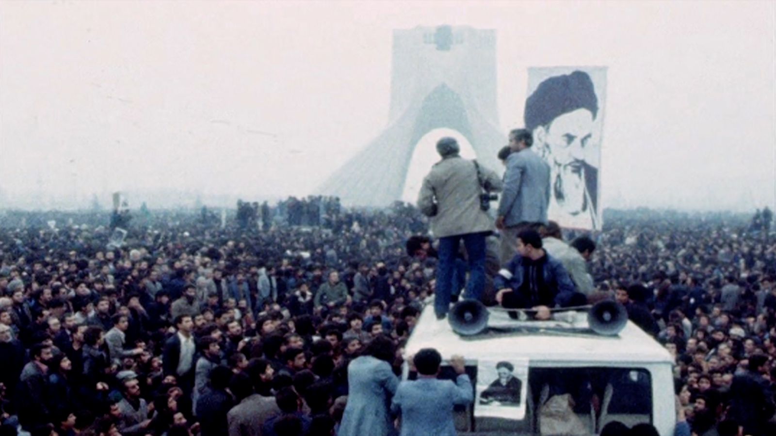 A crowd of people protest in Iran. A few people stand in the foreground, while a large picture of the Shah is in the background.