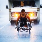 An archival image shows athlete Rick Hansen racing down a rainy highway. There is an RV behind him.