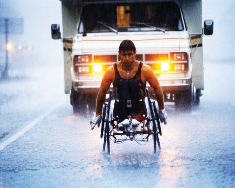 An archival image shows athlete Rick Hansen racing down a rainy highway. There is an RV behind him.