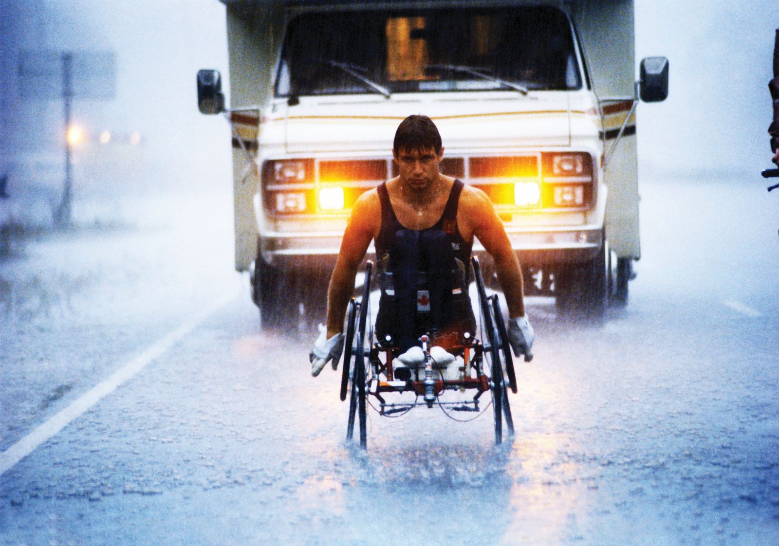 An archival image shows athlete Rick Hansen racing down a rainy highway. There is an RV behind him.