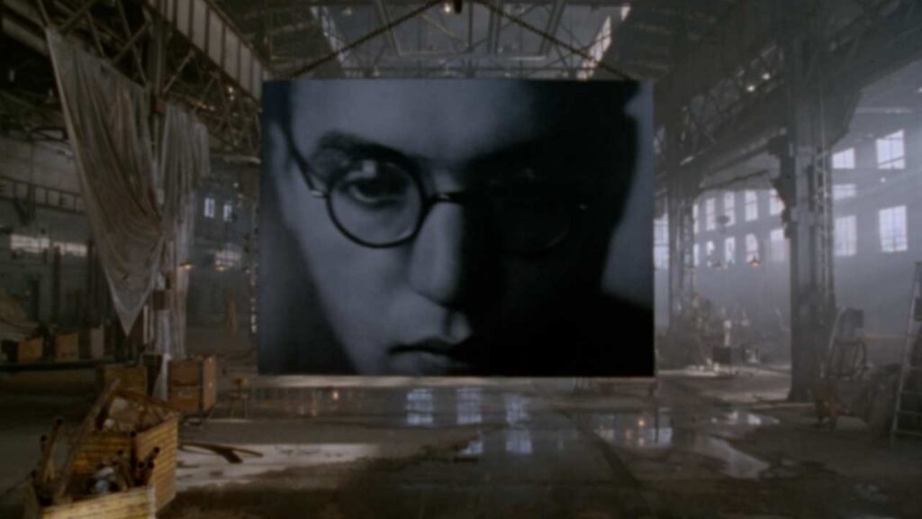 An image of composer Kurt Weill appears on a projection screen that hangs from the ceiling of a large warehouse.