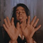 Singer Nick Cave performs in close-up facing the camera. His hands are open-palm forward towards the lens. He has long brown hair and appears in a warehouse.
