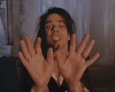 Singer Nick Cave performs in close-up facing the camera. His hands are open-palm forward towards the lens. He has long brown hair and appears in a warehouse.