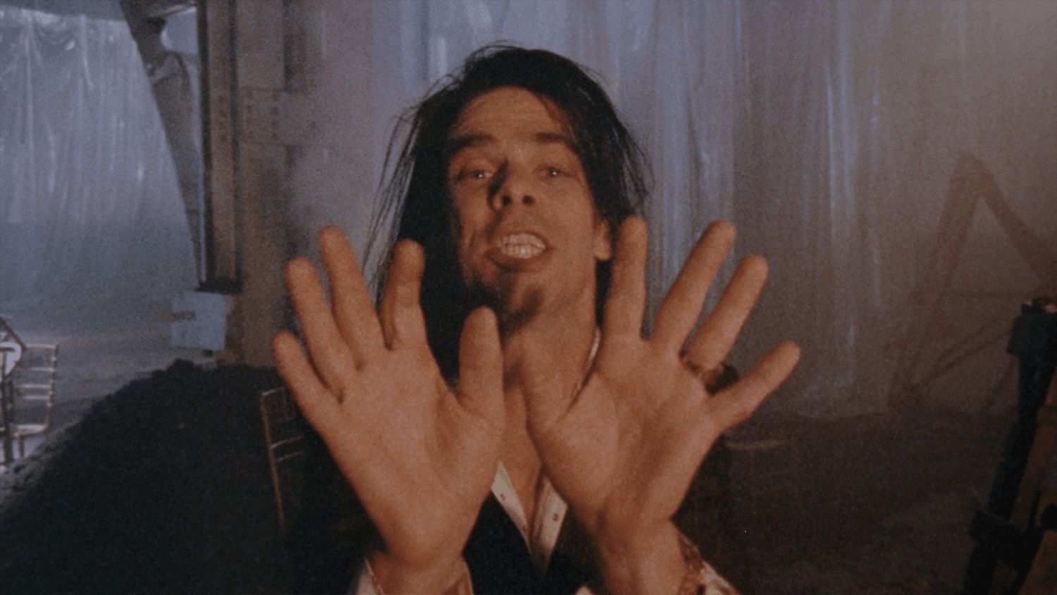 Singer Nick Cave performs in close-up facing the camera. His hands are open-palm forward towards the lens. He has long brown hair and appears in a warehouse.