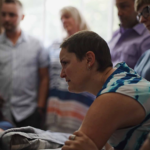 A woman in a white and blue shirt lens over a hospital bed. She had a very serious expression. Many people stand in the background.
