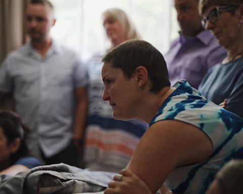 A woman in a white and blue shirt lens over a hospital bed. She had a very serious expression. Many people stand in the background.