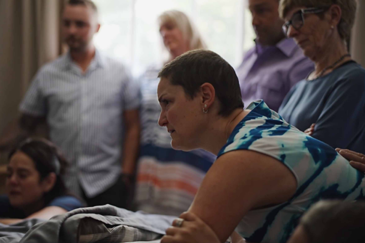 A woman in a white and blue shirt lens over a hospital bed. She had a very serious expression. Many people stand in the background.