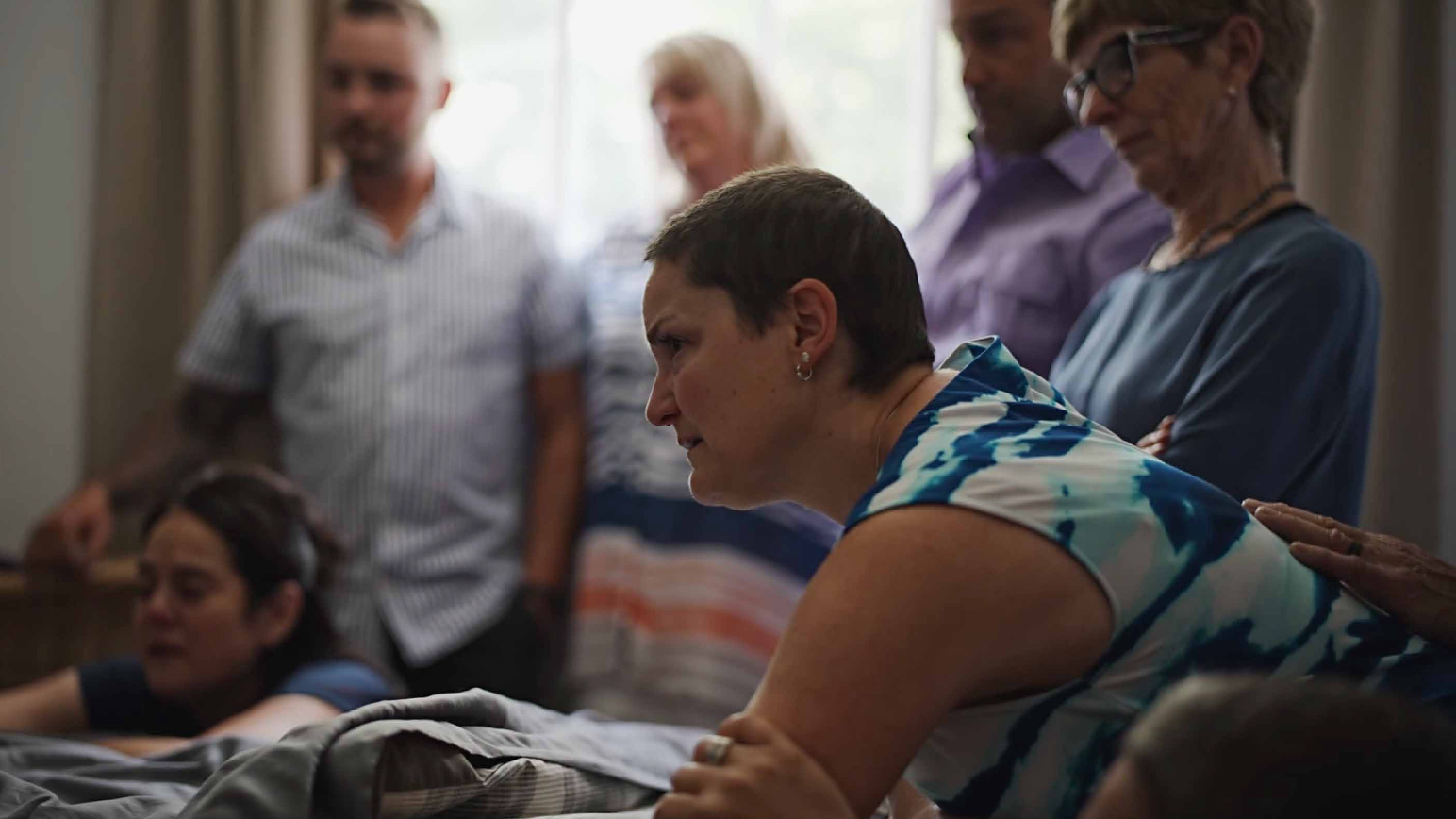 A woman in a white and blue shirt lens over a hospital bed. She had a very serious expression. Many people stand in the background.