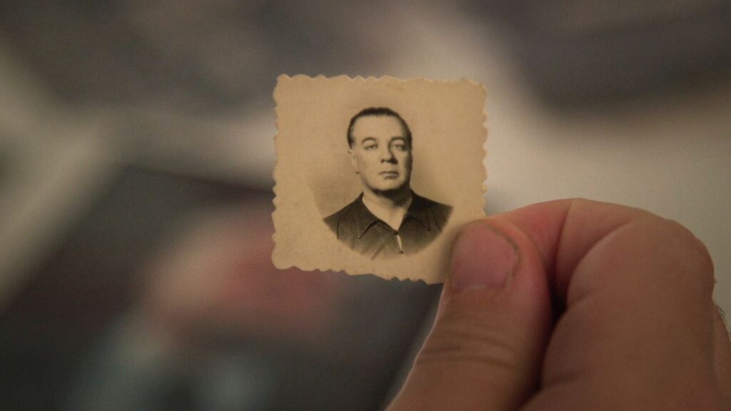 A close-up of a thumb and index finger holding a very small passport-style photo. The photograph is very old and offers a black and white image of a a man with an expressionless face. The man in the photo is of light complexion and wearing a black shirt.