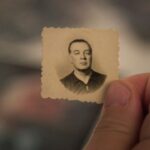 A close-up of a thumb and index finger holding a very small passport-style photo. The photograph is very old and offers a black and white image of a a man with an expressionless face. The man in the photo is of light complexion and wearing a black shirt.