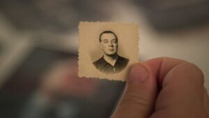 A close-up of a thumb and index finger holding a very small passport-style photo. The photograph is very old and offers a black and white image of a a man with an expressionless face. The man in the photo is of light complexion and wearing a black shirt.