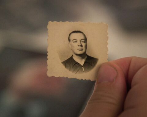 A close-up of a thumb and index finger holding a very small passport-style photo. The photograph is very old and offers a black and white image of a a man with an expressionless face. The man in the photo is of light complexion and wearing a black shirt.