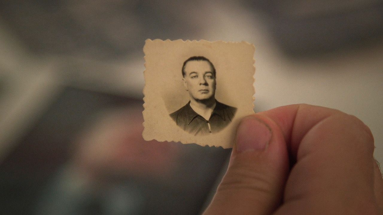 A close-up of a thumb and index finger holding a very small passport-style photo. The photograph is very old and offers a black and white image of a a man with an expressionless face. The man in the photo is of light complexion and wearing a black shirt.