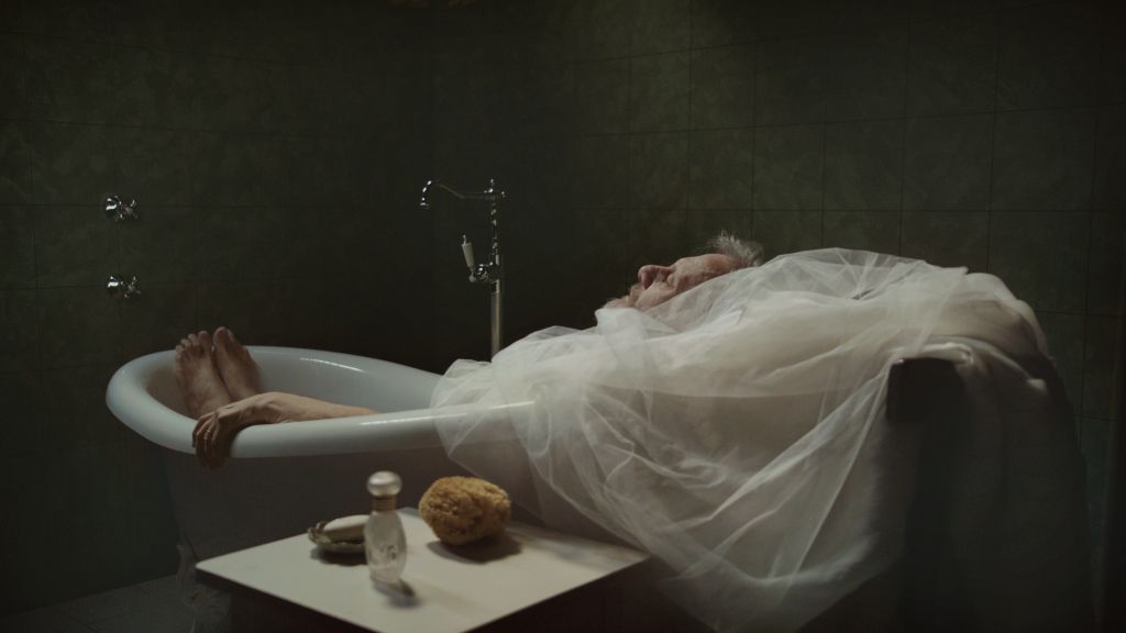 Philosopher Slavoj Zizek reclines in a white bathtub with a wedding dress flipped over his shoulders, mimicking an image from the film Melancholia