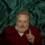 Slovenian philosopher Slavoj Zizek is pictured from above as he lies on a bed with blue satin sheets. He is a white man with grey hair and is wearing a burgundy sweater with a white collared short underneath. His fingered is raised in the air as he makes an intellectual argument.