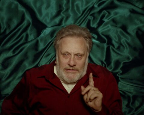 Slovenian philosopher Slavoj Zizek is pictured from above as he lies on a bed with blue satin sheets. He is a white man with grey hair and is wearing a burgundy sweater with a white collared short underneath. His fingered is raised in the air as he makes an intellectual argument.