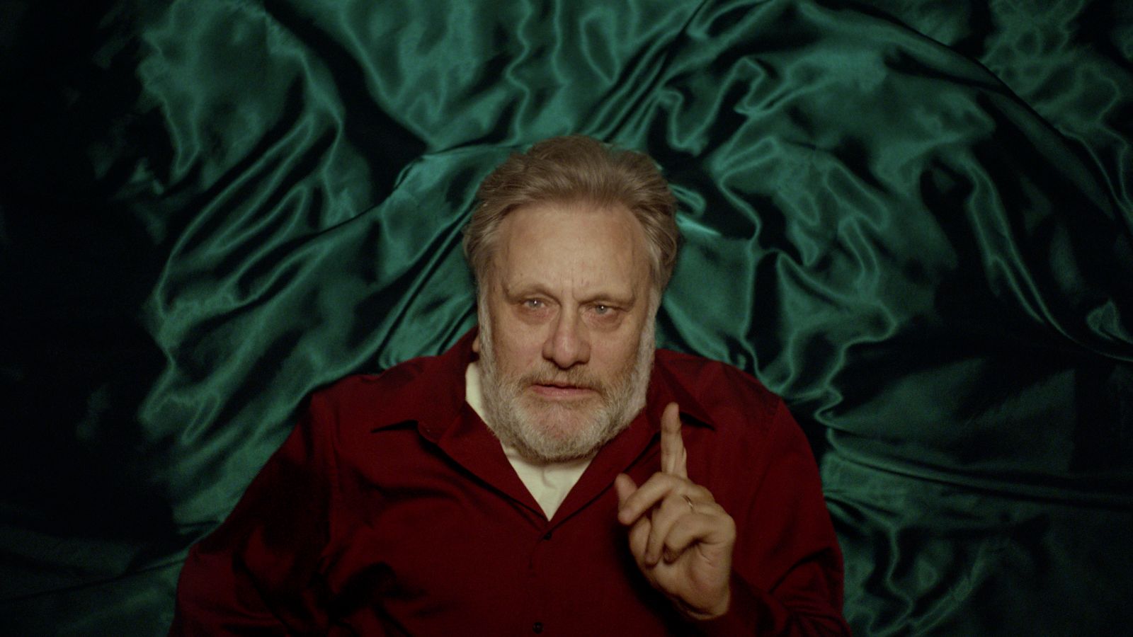 Slovenian philosopher Slavoj Zizek is pictured from above as he lies on a bed with blue satin sheets. He is a white man with grey hair and is wearing a burgundy sweater with a white collared short underneath. His fingered is raised in the air as he makes an intellectual argument.