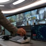 The image shows a large wall of surveillance monitors that capture various aspects of urban life. In the foreground of the picture is the back of a man operating a control panel while facing the wall of monitors. He is wearing a black-sleeved shirt and his hand is manoeuvring a joystick to control the gaze of the monitors.