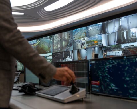 The image shows a large wall of surveillance monitors that capture various aspects of urban life. In the foreground of the picture is the back of a man operating a control panel while facing the wall of monitors. He is wearing a black-sleeved shirt and his hand is manoeuvring a joystick to control the gaze of the monitors.