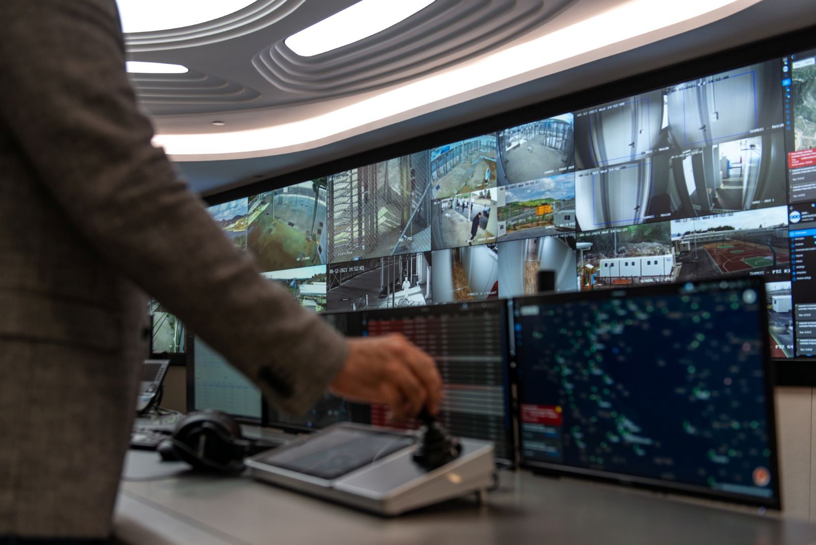 The image shows a large wall of surveillance monitors that capture various aspects of urban life. In the foreground of the picture is the back of a man operating a control panel while facing the wall of monitors. He is wearing a black-sleeved shirt and his hand is manoeuvring a joystick to control the gaze of the monitors.