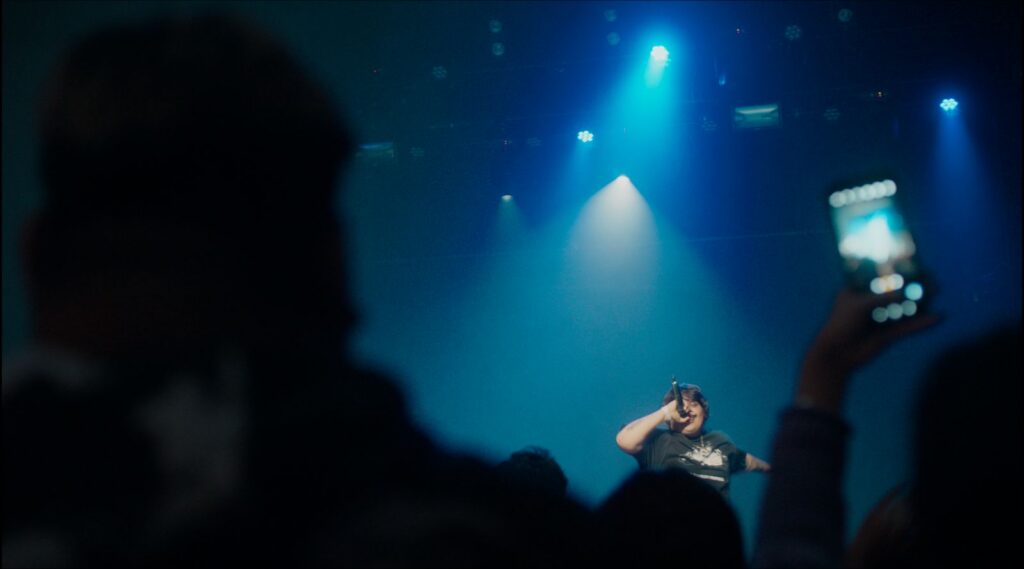 A long shot shows an Indigenous rapper performing on a stage in front of a crowd. The room is dark and there is a spotlight on him.