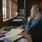 Botanist and author Robin Wall Kimmerer sits at a desk in deep thought while looking out a window. She is wearing a blue sleeved shirt and has long grey hair. Her desk is covered with a mess of papers.