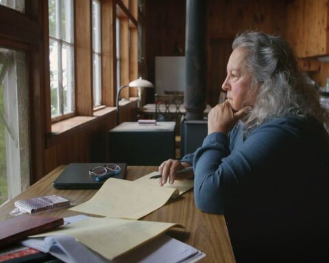 Botanist and author Robin Wall Kimmerer sits at a desk in deep thought while looking out a window. She is wearing a blue sleeved shirt and has long grey hair. Her desk is covered with a mess of papers.