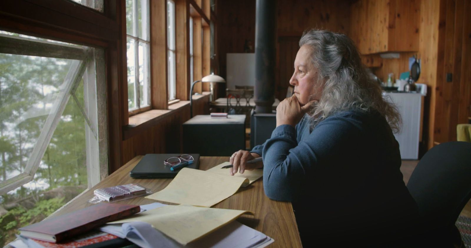 Botanist and author Robin Wall Kimmerer sits at a desk in deep thought while looking out a window. She is wearing a blue sleeved shirt and has long grey hair. Her desk is covered with a mess of papers.