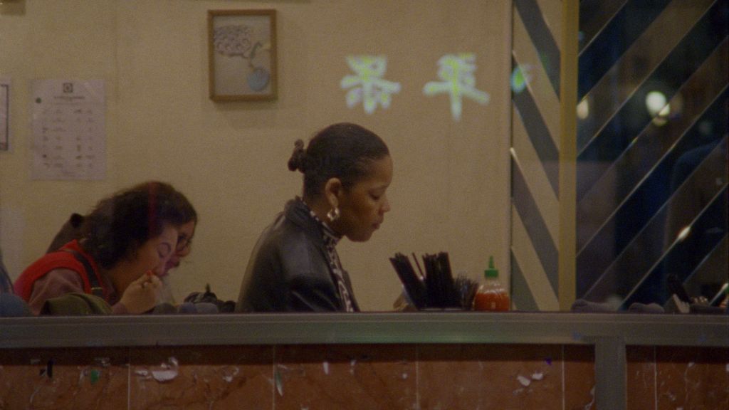 Two women, one Black and one white, sit at desks. They are pictured from a distance, as if someone is looking at them through a window. They are in a setting that resembles an office or classroom.