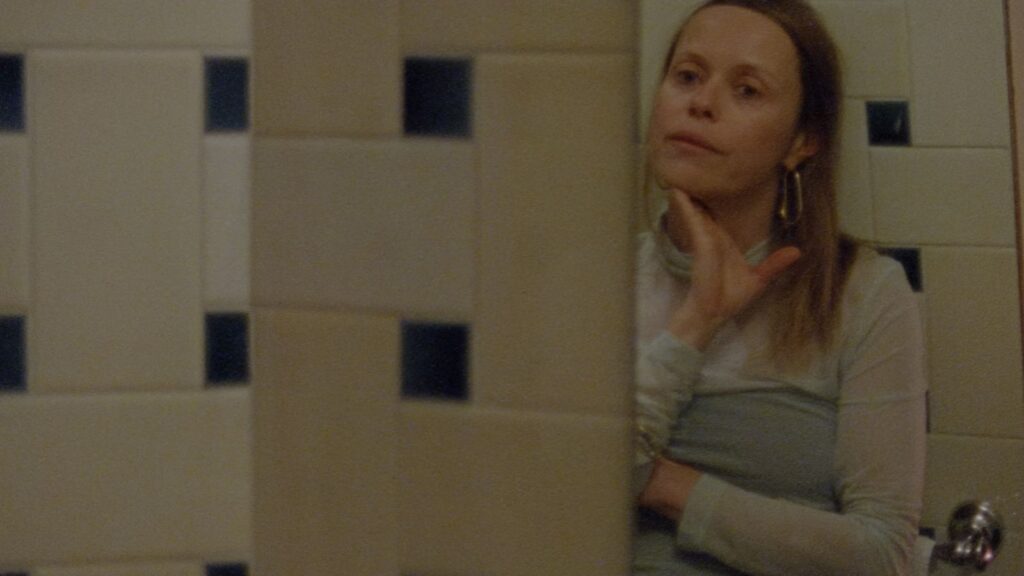 Filmmaker Beatrice Gibson stands looking at her reflection in a mirror in a tiled bathroom. She is a white woman with blonde hair, and is wearing a light blue sleeved dress. Her hand is caressing her chin and she looks to be in deep thought.