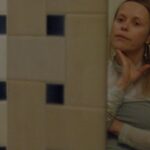 Filmmaker Beatrice Gibson stands looking at her reflection in a mirror in a tiled bathroom. She is a white woman with blonde hair, and is wearing a light blue sleeved dress. Her hand is caressing her chin and she looks to be in deep thought.