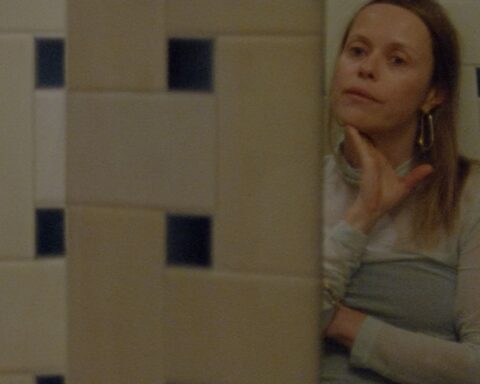 Filmmaker Beatrice Gibson stands looking at her reflection in a mirror in a tiled bathroom. She is a white woman with blonde hair, and is wearing a light blue sleeved dress. Her hand is caressing her chin and she looks to be in deep thought.