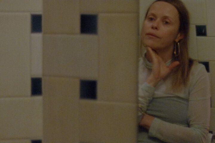 Filmmaker Beatrice Gibson stands looking at her reflection in a mirror in a tiled bathroom. She is a white woman with blonde hair, and is wearing a light blue sleeved dress. Her hand is caressing her chin and she looks to be in deep thought.