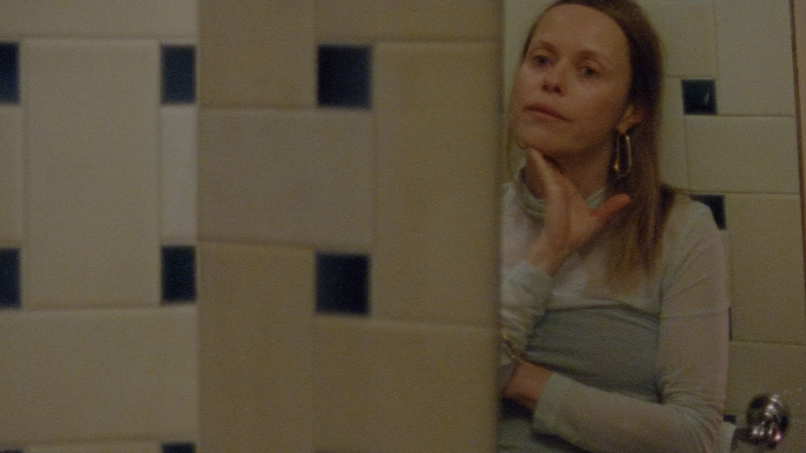 Filmmaker Beatrice Gibson stands looking at her reflection in a mirror in a tiled bathroom. She is a white woman with blonde hair, and is wearing a light blue sleeved dress. Her hand is caressing her chin and she looks to be in deep thought.
