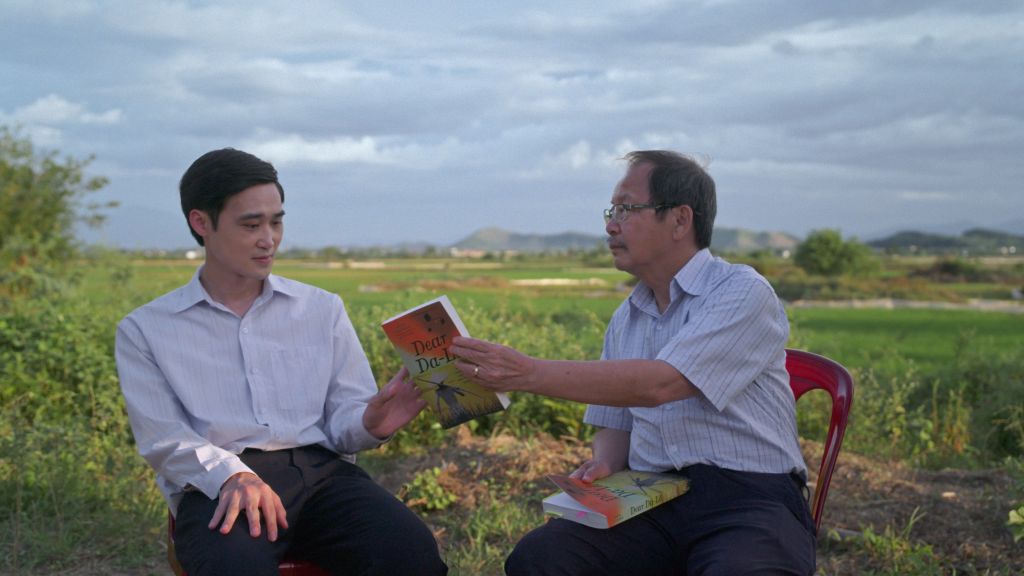 Two Vietnamese men sit in a field. The older man seated on the right hands a book to the younger man seated on the left. They are both wearing white collared shirts, and it is an overcast day.