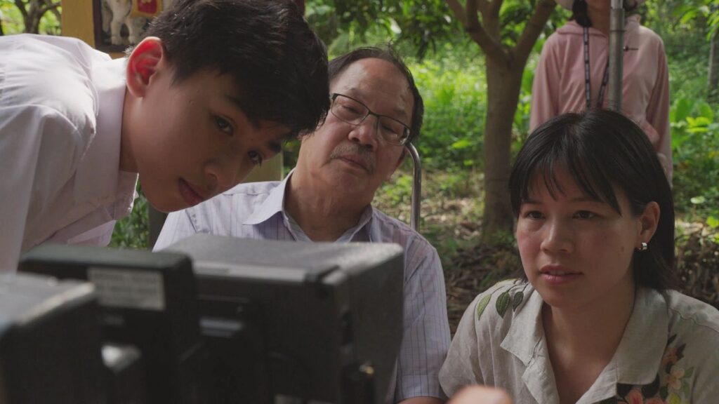 Vietnamese-Canadian filmmaker Ashley Duong and her father, along with the actor portraying him as a young boy, watch a monitor while on location. They are seated in a lush green area with many trees.
