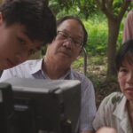 Vietnamese-Canadian filmmaker Ashley Duong and her father, along with the actor portraying him as a young boy, watch a monitor while on location. They are seated in a lush green area with many trees.