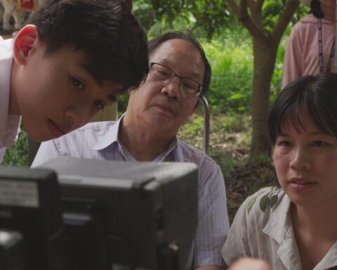 Vietnamese-Canadian filmmaker Ashley Duong and her father, along with the actor portraying him as a young boy, watch a monitor while on location. They are seated in a lush green area with many trees.