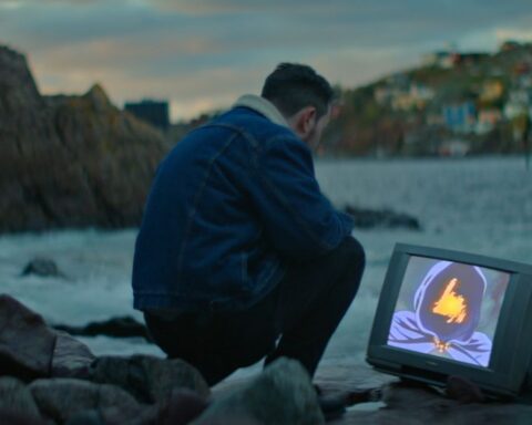 A man crouches down on a rocky shore in Newfoundland, looking at a TV that is positioned on the ground by the water. There is a strange image on the screen with a figure in a dark purple hood and an orange light where his face should be.