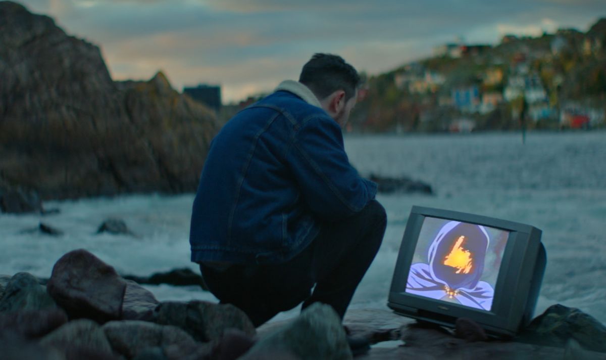 A man crouches down on a rocky shore in Newfoundland, looking at a TV that is positioned on the ground by the water. There is a strange image on the screen with a figure in a dark purple hood and an orange light where his face should be.